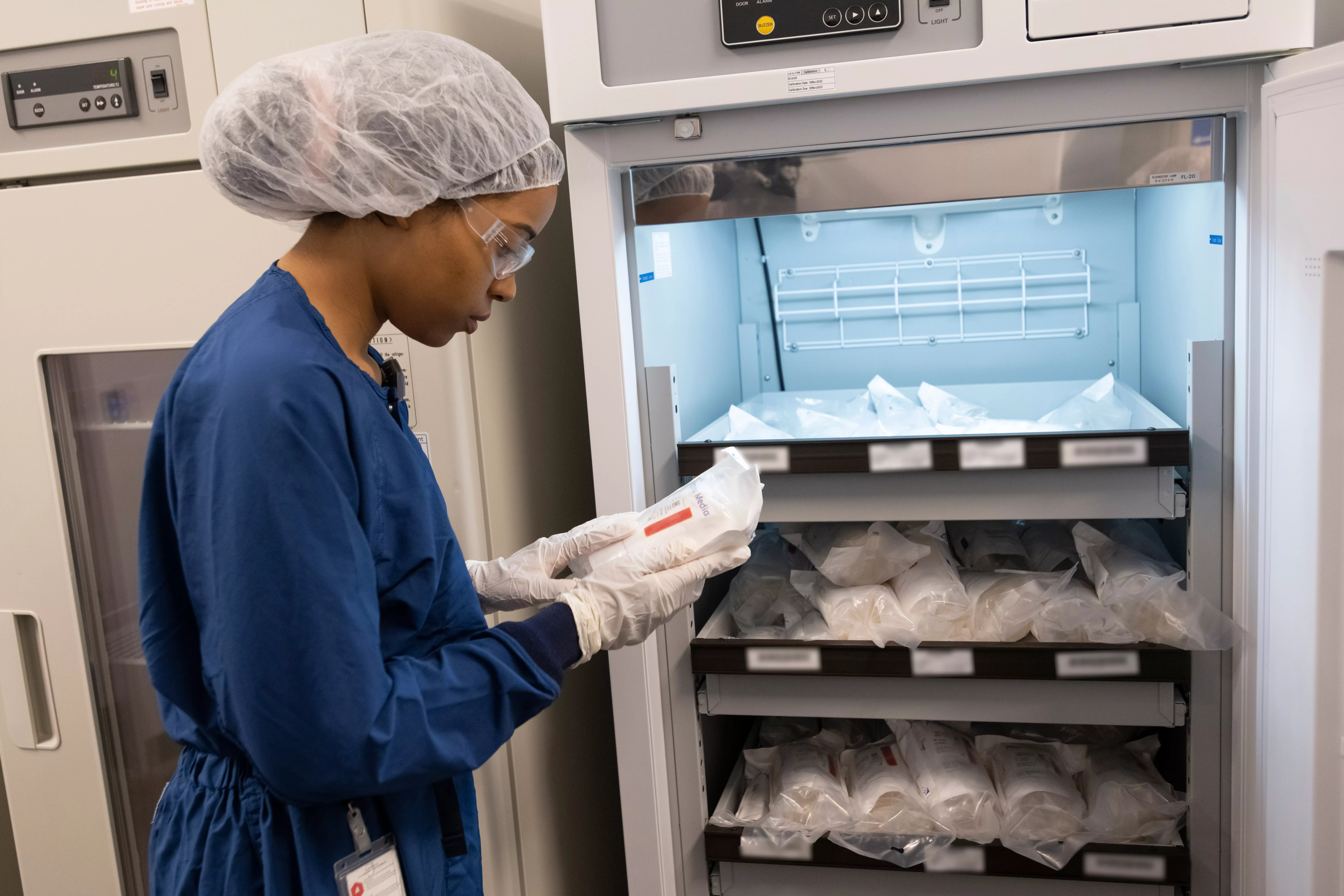 An associate in personal protective equipment stands at a refrigerator examining a bag An associate in personal protective equipment stands at a refrigerator examining a bag